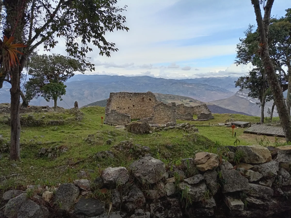 Los muros curvos de piedra parcialmente en pie de una vivienda circular en ruinas en Kuélap, que alcanzan aproximadamente la altura de una persona en una colina cubierta de hierba, con las montañas de Amazonas al fondo