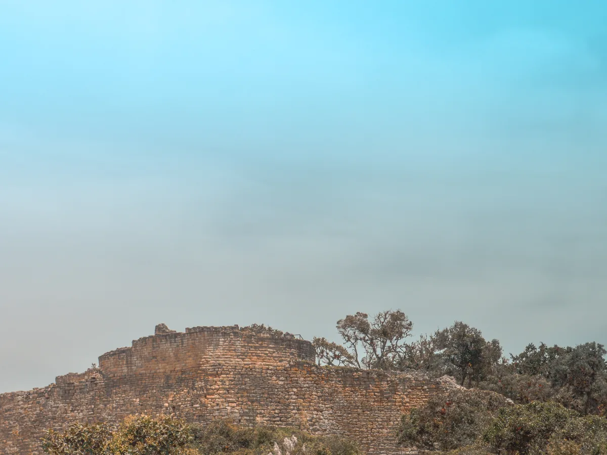 La muralla perimetral de Kuélap vista desde la distancia a lo largo de la cresta del bosque de niebla, con bajos parapetos de piedra recortados contra el cielo y árboles creciendo en la línea de la cima